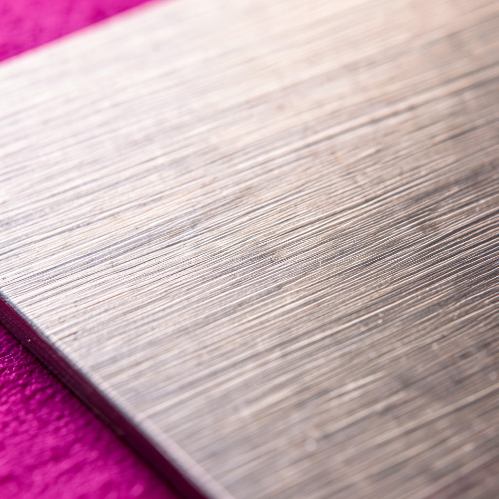 A macro photograph of brushed metal surface in warm silver-gray, with fine horizontal grain lines catching soft light.